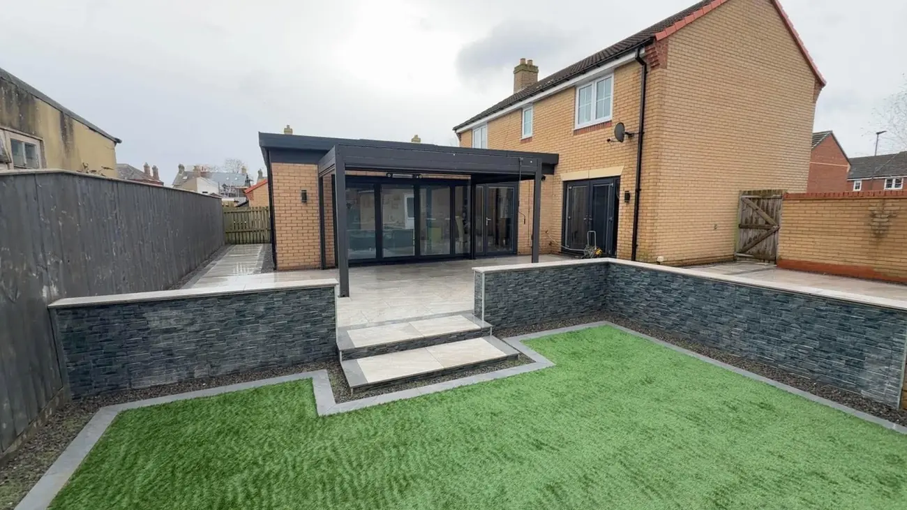 Stone-effect clad raised walls with steps, porcelain paving, artificial grass and a pergola